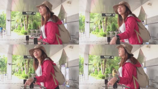 美丽的亚洲女人，一个长长的黑发，帽子，肩包和行李在手推车上等待机场候机楼寻找出租车，交通工具探索新的旅程。高清在线视频素材下载