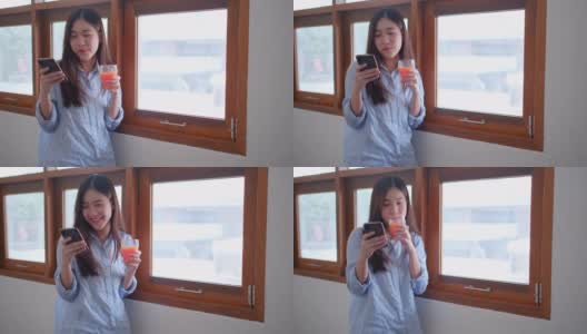 漂亮的女孩或女人在玻璃窗前用手机和喝橙汁，看起来在家里很开心高清在线视频素材下载