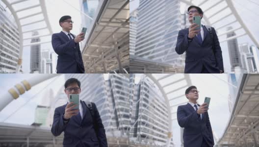 自信的亚洲商人，西装革履的办公室职员在城市中行走，用智能手机在线发短信。办公区内使用手机进行网上办公的商人高清在线视频素材下载