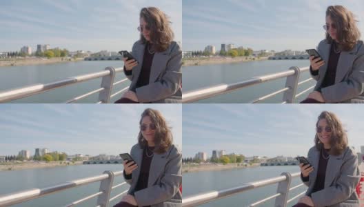 一名女子坐在城市的河边，用她的智能手机在室外打字高清在线视频素材下载