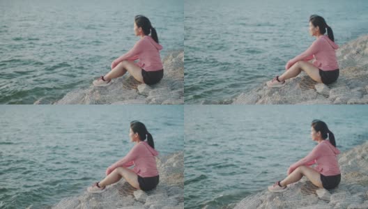 年轻女子跑步者坐在海边的石头上，从锻炼中休息，欣赏海滩日落的景色。高清在线视频素材下载