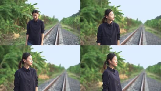 一个年轻美丽的亚洲女人，她把头发绑在后面，穿着黑色的衬衫和牛仔裤，走着，想着她在路边有香蕉树森林的地方铁路上的生活，然后她停下来向前看。高清在线视频素材下载