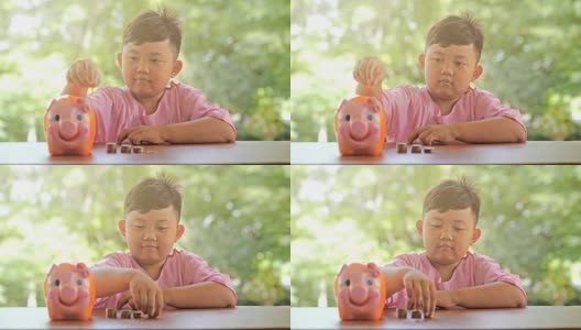 亚洲男孩带着快乐的微笑把硬币放入储蓄罐，慢镜头。散景背景。存钱，为将来攒钱。高清在线视频素材下载