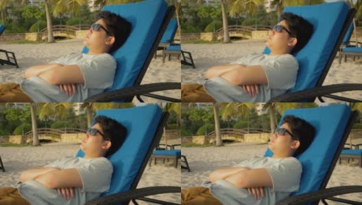 快乐的亚洲男孩戴着太阳镜在沙滩椅上日光浴在早上度假期间在酒店或度假村与家人，生活方式的概念。高清在线视频素材下载