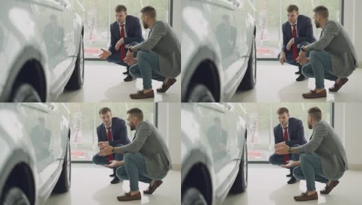 两名兴高采烈的顾客和友好的推销员蹲在一辆昂贵的汽车旁，一边交谈一边打手势讨论汽车型号。购买和销售汽车的概念。高清在线视频素材下载