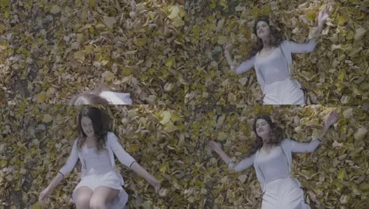 迷人的年轻女子穿着白色的连衣裙，微笑和放松躺在地上的秋天的树叶慢动作高清在线视频素材下载