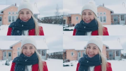 一个年轻的金发女人的肖像在红色大衣与围巾微笑进入相机，而站在飘落的雪。在后院田园诗般的房子与冬天的景色。高清在线视频素材下载