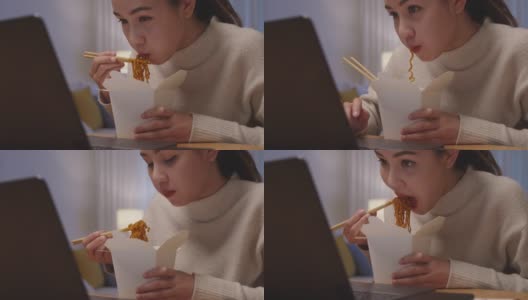 年轻迷人美丽的亚洲女性饿吃方便面中国食品盒带走满嘴看电脑笔记本在家里在忙工作从家里深夜多任务不健康的饮食。高清在线视频素材下载