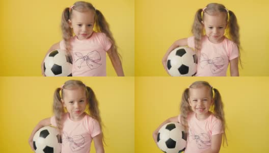 真实可爱的微笑学龄前小女孩与经典的黑白足球看着摄像机在黄色背景。孩子们穿着t恤和短裤踢足球。运动，冠军，团队概念高清在线视频素材下载