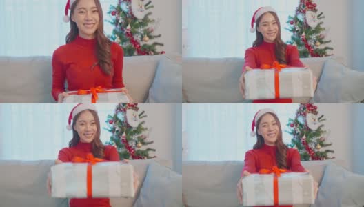 亚洲美丽的年轻女人的肖像持有圣诞礼物盒。漂亮的女孩穿着红布衣服戴着圣诞老人的帽子坐在沙发上，准备在家里庆祝节日圣诞节并微笑着看着摄像机。高清在线视频素材下载