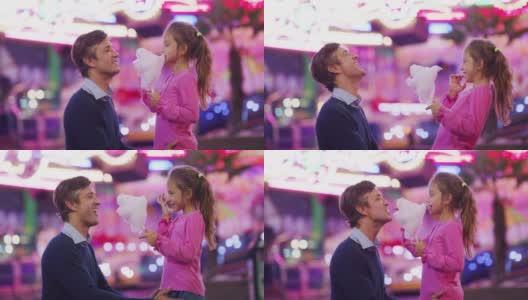 真实的照片，一个快乐的父亲和他的小女儿正在开心地吃棉花糖在游乐园与月亮公园的灯光在晚上。高清在线视频素材下载
