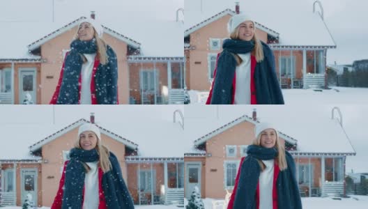 一个年轻的金发女人的肖像在红色大衣与围巾在她的后院行走和跳跃，而雪花落。背景是田园诗般的房子，带有冬季景色。高清在线视频素材下载