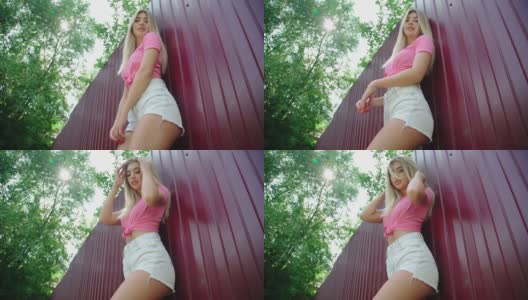 一位欢快的金发年轻女子身穿粉色背心和牛仔短裤，在深红色的铁栅栏前摆姿势。积极的情绪。暑假。低的观点。人们生活方式的概念。高清在线视频素材下载