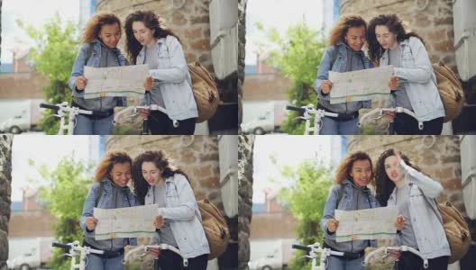 两个游客站在街上，手里拿着自行车，一边看地图一边聊天。迷人的年轻女子和休闲服装与背包，笑着环顾四周。高清在线视频素材下载