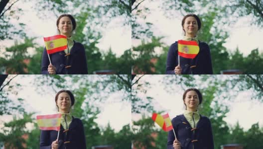 慢动作肖像，漂亮的年轻女子西班牙球迷挥舞西班牙官方旗帜，看着镜头与美好的微笑。背景是绿树和天空。高清在线视频素材下载
