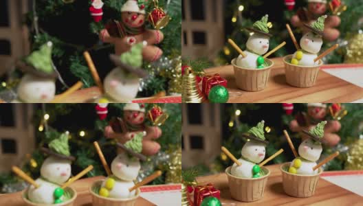 照片:在一间装饰着圣诞树的公寓里，有两个自制的冰霜雪人杯形蛋糕，用冰霜球做的，黑巧克力饼干，绿色奶油做的帽子，粉色的鼻子和巧克力球做的绿色纽扣，椒盐卷饼棒做的手臂高清在线视频素材下载