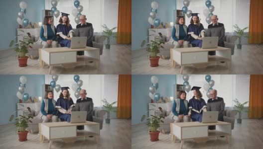 毕业典礼，欢乐的家庭和女儿在学术服装庆祝在线毕业典礼，而坐在家里的沙发上气球的背景关于社会距离的时间高清在线视频素材下载