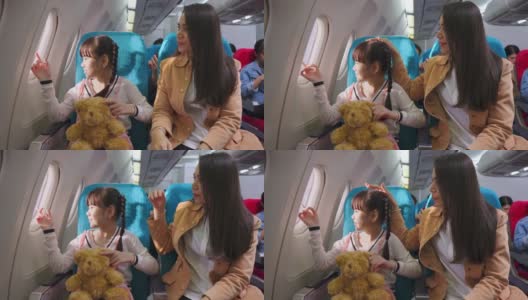 亚洲母女坐在飞机上，一起望着窗外。当飞机在空中飞行时，抱着熊娃娃的女孩在看。航空运输的概念。高清在线视频素材下载