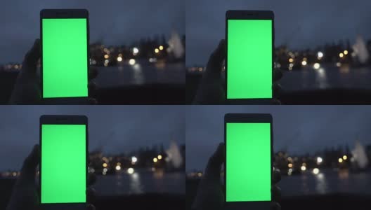 男子手握白色智能手机，在夜城灯光散景的背景下。绿色的屏幕。高清在线视频素材下载