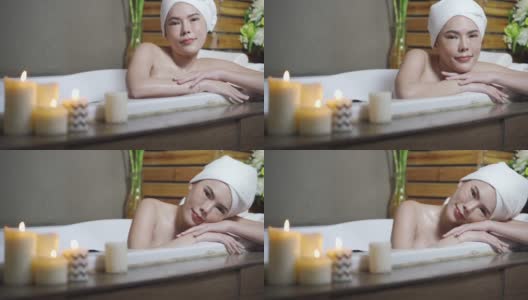 浴缸里点着蜡烛的性感女人。放松的女人泡在肥皂浴里，在装饰着蜡烛的健康水疗概念的按摩浴缸边睡觉。高清在线视频素材下载