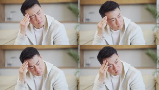 一个年轻的亚洲男人在家里的沙发上抱着他严重头痛的头高清在线视频素材下载