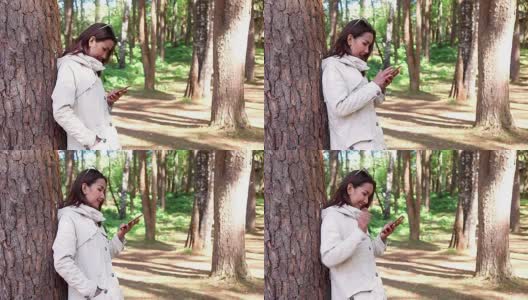 年轻美丽的女人Lean the Trees和她的手机聊天穿长外套夹克在寒冷的天气在国家松树森林公园与幸福。高清在线视频素材下载