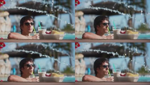 年轻英俊的男人戴着墨镜，喝着鸡尾酒，吃着热带水果，在旅游度假的游泳池里放松。1920 x1080高清在线视频素材下载