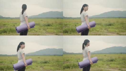 迷人的亚洲女人站着，握着瑜伽垫在美丽的山景在早上。高清在线视频素材下载