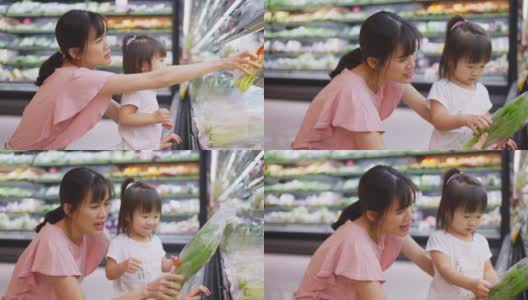 年轻的亚洲美丽的母亲拿着购物篮和她的孩子走在超市。她从货架上挑选绿色沙拉蔬菜，然后给她的小孩吃。健康的购物。高清在线视频素材下载