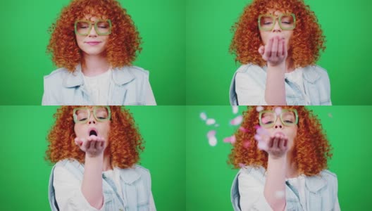 年轻俏皮的红发卷发女人向相机吹五彩纸屑，微笑着越过绿色的色度键背景高清在线视频素材下载