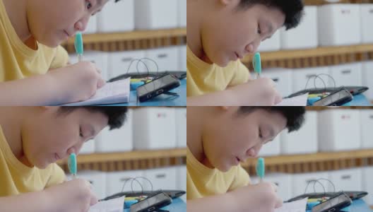 亚洲男孩一边做作业一边在家学习在线课程，社交距离的生活方式概念。高清在线视频素材下载