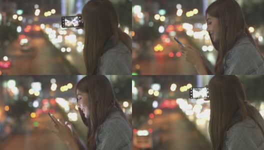 微笑的年轻美丽的亚洲女人用智能手机上网社交媒体或网上购物，并拍下城市的路灯。快乐的漂亮女孩喜欢城市夜生活。高清在线视频素材下载