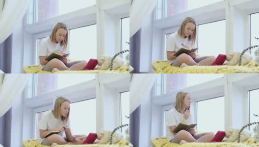 十几岁的女孩坐在窗边的窗台上，在日记里画画或写东西。可爱的女孩在平板电脑上看视频高清在线视频素材下载