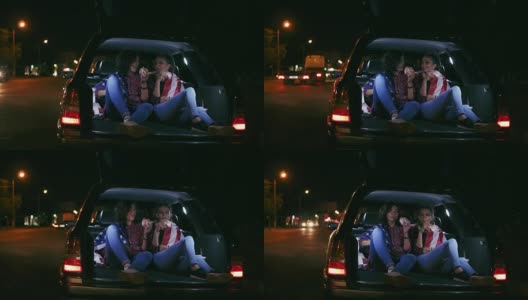 两个迷人的女朋友坐在敞开的后备箱，在城市的夜晚聊天和吃汉堡。Slowmotion拍摄高清在线视频素材下载