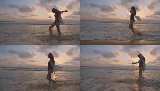 快乐的女人穿着泳衣和衬衫在日落时走在海滩上。海浪拍打着女性的脚。年轻美丽的女孩享受生活，在海边玩得开心。暑假。慢动作高清在线视频素材下载