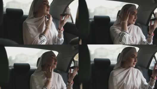 一个年轻的穆斯林妇女在米色头巾的肖像，坐在车里，看着一个小化妆镜，检查她的化妆高清在线视频素材下载