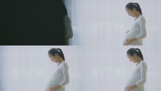 美丽的亚洲孕妇透过窗户看，并在客厅揉胃。高清在线视频素材下载