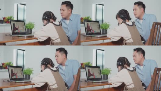 由于冠状病毒大流行，亚洲小女孩通过数字远程互联网会议从学校老师那里学习在线课程。孩子看着电脑笔记本电脑屏幕坐在父亲。高清在线视频素材下载