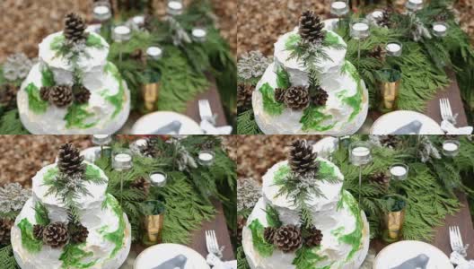 这是一个两层楼的白色婚礼蛋糕的特写镜头，上面装饰着松果和云杉树枝，上面的桌子上摆满了用蜡烛装饰的盘子和玻璃杯。高清在线视频素材下载
