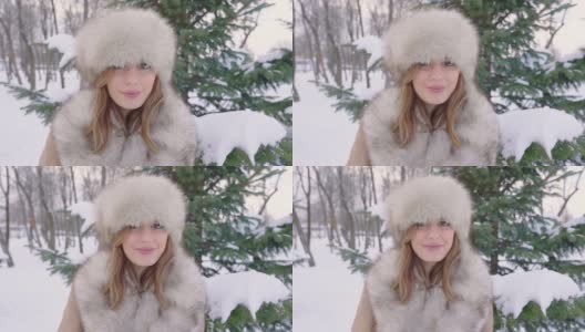 美丽的女人戴着皮帽看着相机在冬天的城市公园里白天在下雪的天气下飘落雪花，慢镜头高清在线视频素材下载