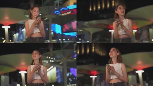 年轻亚洲女人的4K肖像走在城市的街道听音乐从耳机在晚上高清在线视频素材下载