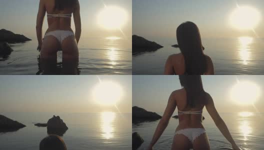 年轻女子在弦游泳衣触摸海水在日出的背景。缓慢的运动。女孩站在热带海滩上，穿着白色比基尼享受夏天的海洋高清在线视频素材下载