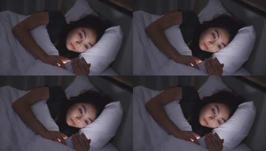 一名亚洲年轻女子晚上躺在床上玩智能手机，在卧室熄灯。在手机上阅读社交。在昏暗的光线下使用手机对眼睛有健康影响。失眠的概念高清在线视频素材下载