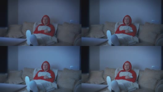一个穿着连帽运动衫的男人，晚上坐在沙发上，把脚放在前面看电视，电视发出的光高清在线视频素材下载