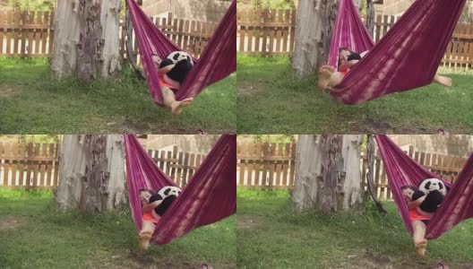 年轻快乐的小女孩在夏令营躺在彩色的吊床上放松地玩在外面的毛绒狗娃娃在绿色的草地上孩子们，孩子们的慢动作高清在线视频素材下载