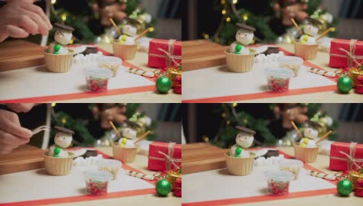 在装饰着圣诞树和装饰品的厨房里，准备冬天传统节日圣诞节的烘焙食品和甜点架，用糖霜自制纸杯蛋糕做一个雪人，用镊子给眼睛加巧克力高清在线视频素材下载