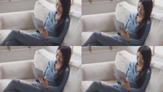 美丽的肖像年轻的亚洲女人微笑和快乐的使用数字平板电脑坐在客厅的沙发上，女孩与满足社交网络在线，商业交流的概念。高清在线视频素材下载
