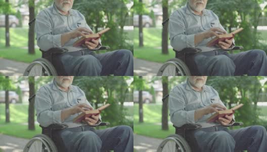 无法辨认的瘫痪老人翻动书页，大声朗读。白发苍苍的退休白人在阳光明媚的夏日公园里享受文学。爱好和残疾。高清在线视频素材下载