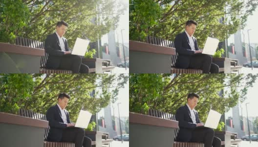 年轻的亚洲商人或自由职业者坐在长椅和工作笔记本电脑在城市公园在现代城市街道高清在线视频素材下载