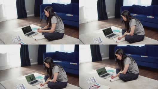 从侧面看亚洲女学生坐在家里的地板上，在线学习，观看与老师的视频会议，并在笔记本上做笔记高清在线视频素材下载